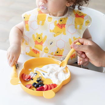 A child enjoys yogurt with fruit on a bear plate, using Bumkins Winnie the Pooh Silicone Dipping Spoons with adult assistance.