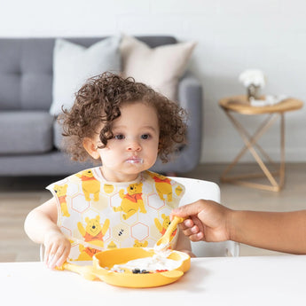 A curly-haired toddler joyfully self-feeds with a Bumkins Winnie the Pooh silicone dipping spoon, food adorably smudged on their cheeks.