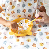 A child self-feeds with a Bumkins Winnie the Pooh silicone spoon from a yellow bear plate, wearing an animal-print shirt.