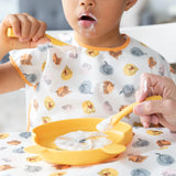 A young child is using a Bumkins Silicone Dipping Spoon: Winnie the Pooh in yellow, with food around their mouth. Theyre holding a matching fork over a yellow plate, while wearing an animal print bib. An adult assists them at the patterned tablecloth, creating an adorable feeding scene.