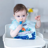 Baby in a high chair, wearing a blue bib with Hangry and dinosaur design, holding a Bumkins Silicone Dipping Spoon: Gumdrop 3 Pack.