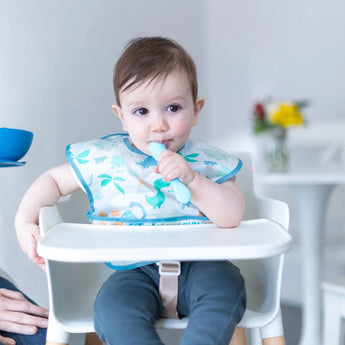 Baby in a highchair with dinosaur bib, holding Bumkins Silicone Dipping Spoon from Gumdrop 3 Pack in bright room with flowers.