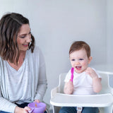 A woman smiles at a baby using Bumkins Lollipop Silicone Spoon, with a purple bowl nearby.