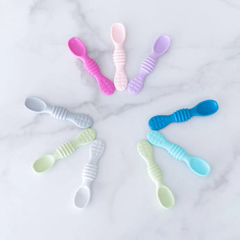 Bumkins Silicone Dipping Spoons in pink, purple, and blue hues for self-feeding are artfully displayed on marble.