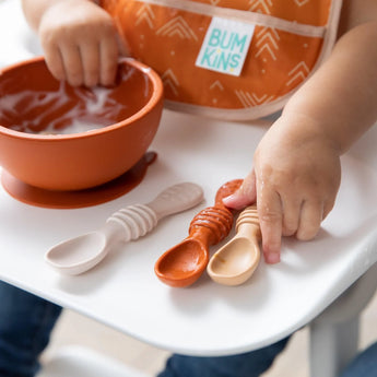 A toddler in a bib sits at a highchair, joyfully exploring self-feeding with Bumkins Silicone Dipping Spoons 3 Pack: Rocky Road.