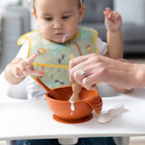 A baby in a bib grips Bumkins Rocky Road Silicone Dipping Spoons in a high chair, with an adult assisting over an orange bowl.