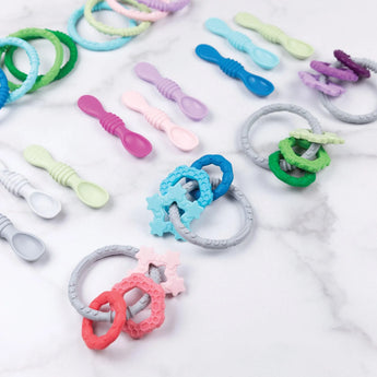 Bumkins Silicone Teething Charms in purple and colorful spoons on marble; pink, blue, green rings, pastel spoons ease teething.