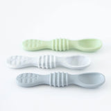 Three pastel silicone dipping spoons from the Bumkins Silicone Dipping Spoons 3 Pack: Taffy are arranged on a white surface. The set includes green, white, and blue spoons with textured grips and rounded bowls, ideal for teaching infants self-feeding skills.