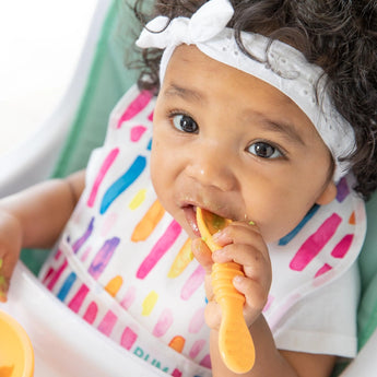 A baby with curls, wearing a white headband and colorful bib, uses a Bumkins Tutti-Frutti Silicone Dipping Spoon adorably.