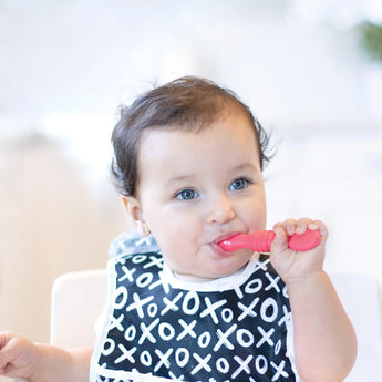 A curly-haired baby in a black bib with white Xs and Os chews on a pink Bumkins Silicone Dipping Spoon in the high chair.