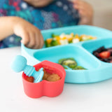 A child explores baby-led weaning with colorful foods on a blue plate, using Bumkins Silicone Chewtensils® in a heart-shaped dish.