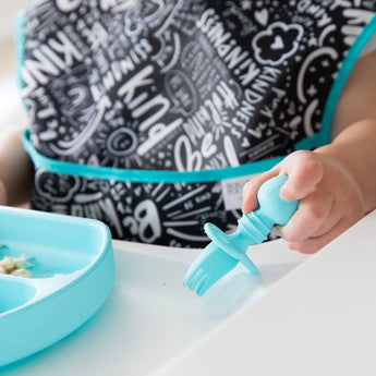 A baby uses Bumkins Silicone Chewtensils®: Blue with a divided tray, their bib showcasing inspiring words.