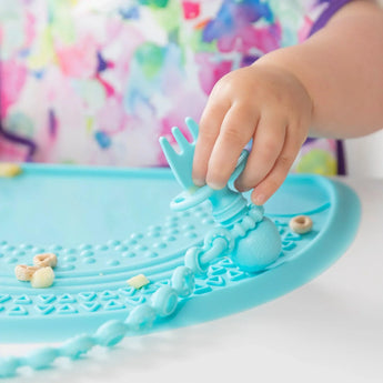 A childs hand explores Bumkins Silicone Chewtensils®: Blue on a mat, with soft utensils among small cereal pieces.