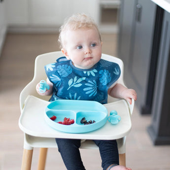 Curly-haired baby in high chair, blue bib on, explores baby-led weaning with Bumkins Silicone Chewtensils® and a toy on the tray.