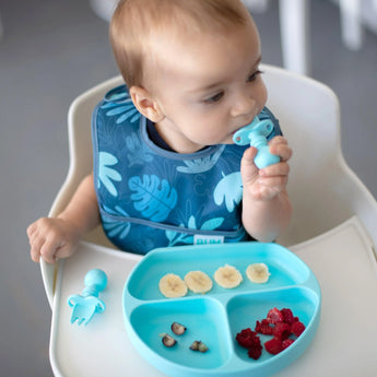 A baby in a high chair uses Bumkins Silicone Chewtensils®: Blue with a sectioned plate of bananas, nuts, and raspberries.