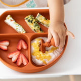 A childs hand reaches for snacks on a Bumkins Silicone Chewtensils® plate with yogurt, fruit, and veggie slices.