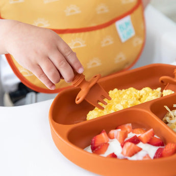 A child uses Bumkins Silicone Chewtensils®: Clay to pick up scrambled eggs from a divided orange plate, with strawberries and yogurt nearby.