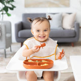 A toddler with pigtails in a high chair uses Bumkins Silicone Chewtensils®, wears a bib, and eats from a divided plate.