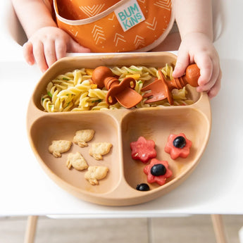 A baby with a Bumkins orange Silicone Chewtensils®: Clay fork reaches over a plate with pasta, fruit flowers, and animal crackers.