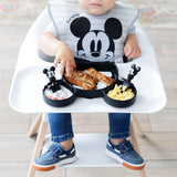 A baby in a high chair enjoys toast, eggs, and fruit using Bumkins Silicone Chewtensils®: Mickey Mouse and a Mickey-themed plate.