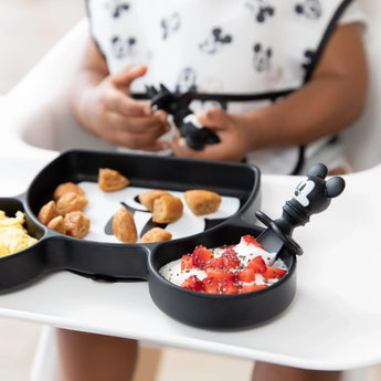 A child holds Bumkins Mickey Mouse Chewtensils with a matching plate of strawberries, pastries, and eggs.