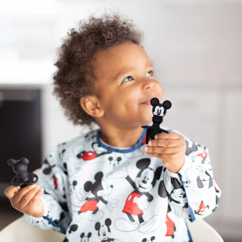 A child smiles in a Bumkins Mickey Mouse outfit, holding Silicone Chewtensils®, perfect for baby-led weaning adventures.