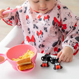 Toddler eating eggs with Bumkins Minnie Mouse Silicone Chewtensils®, playing with Minnie toys, and having toast from a pink bowl.