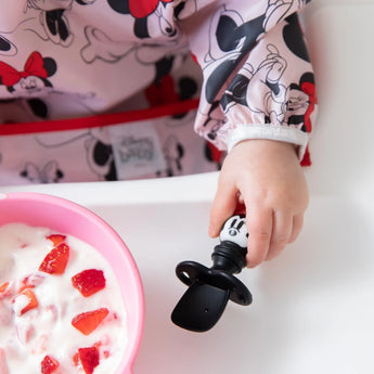 A child in a Disney outfit uses Bumkins Silicone Chewtensils®: Minnie Mouse to enjoy yogurt and strawberries, perfect for baby-led weaning.