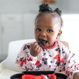 A toddler in a Minnie Mouse bib is happily chewing on Bumkins Silicone Chewtensils®: Minnie Mouse in a high chair.