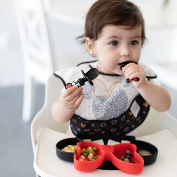 A toddler in a high chair enjoys Minnie Mouse Silicone Chewtensils® by Bumkins on a red Mickey plate, wearing a patterned bib.