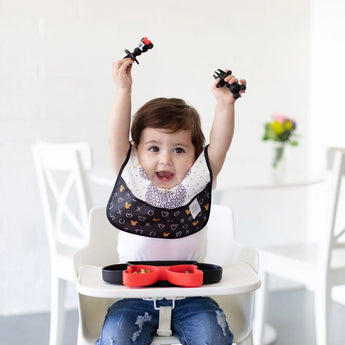 A baby happily raises arms, wearing a bib and holding Bumkins Silicone Chewtensils: Minnie Mouse, with snacks on a red tray nearby.