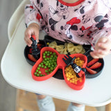 A child in a Minnie Mouse outfit enjoys peas, corn, strawberries, and pasta from a divided tray with Bumkins Silicone Chewtensils®.