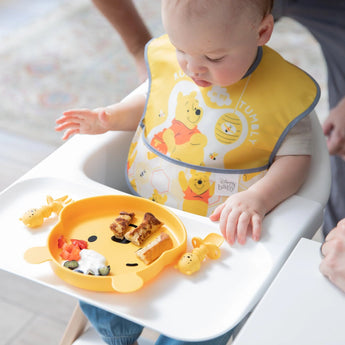 Baby in a high chair wearing a yellow Pooh bib, enjoying toast, berries, and yogurt from a bear plate with Bumkins Silicone Chewtensils.