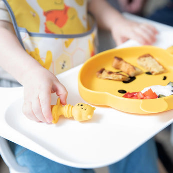 Child in a high chair with Bumkins Silicone Chewtensils®: Winnie The Pooh, enjoying baby-led weaning with toast and tomatoes.