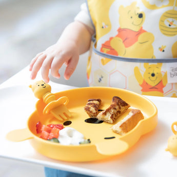 A baby uses Silicone Chewtensils®: Winnie The Pooh by Bumkins to reach for toast, strawberries, and yogurt on a Pooh plate, showing weaning skills.