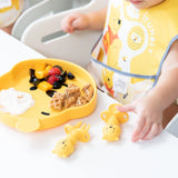 A child in a high chair with a bear bib uses Bumkins Silicone Chewtensils®: Winnie The Pooh, surrounded by toys and waffles, yogurt, and fruit.