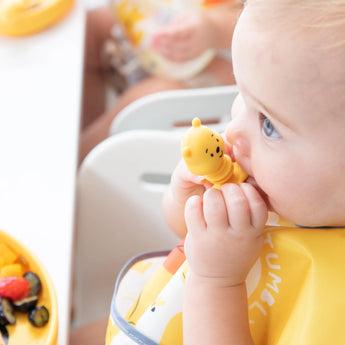 Baby in yellow bib with Bumkins Winnie The Pooh teether, sitting with fruit on a yellow plate, exploring baby-led weaning.