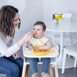 Woman feeds baby in a high chair wearing yellow bib with Bumkins Silicone Chewtensils®: Winnie The Pooh; a table with flowers behind.