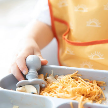 A childs hand uses Bumkins Silicone Chewtensils®: Gray to eat spaghetti from a tray, wearing a yellow sun-patterned bib.