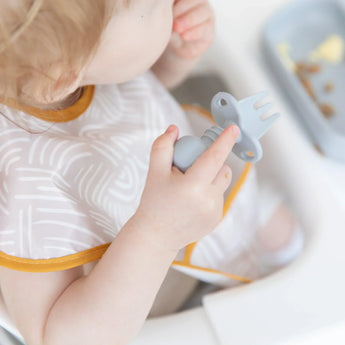 A toddler in a high chair, wearing a bib, holds Bumkins Silicone Chewtensils®: Gray fork, enjoying baby-led weaning with small food pieces.