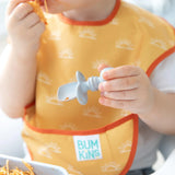 A toddler joyfully embraces baby-led weaning with Bumkins yellow bib featuring sun patterns. Clutching a Silicone Chewtensils®: Gray spoon, spaghetti is near their mouth and in a bowl beside them, turning mealtime into an enjoyable adventure.