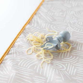 A Bumkins Silicone Chewtensils®: Gray pacifier lies in tangled spaghetti on a patterned surface with an orange border.