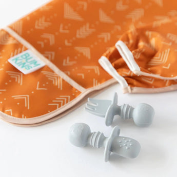 Bumkins Orange bib & two gray Silicone Chewtensils®, ideal for baby-led weaning, on a white surface.