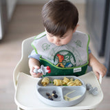 A toddler in a highchair enjoys baby-led weaning with pasta on a food-grade silicone plate, wearing a bib and holding Bumkins Silicone Chewtensils®.