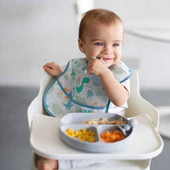 A baby using Bumkins Silicone Chewtensils explores BLW with corn, chicken, and carrots in a high chair, wearing a colorful bib.