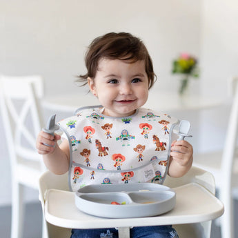 Toddler grins in a high chair, wearing a bib and holding Bumkins Silicone Chewtensils®: Gray over a plate with food.