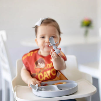 Baby in high chair, red bib, holding Bumkins Silicone Chewtensils® in gray with a white plate. Blurry background.