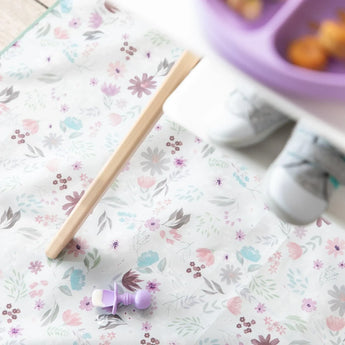 Floral baby mat with Bumkins Lavender Silicone Chewtensils®. Childs shoes blurred above a baby-led weaning meal on a tray.