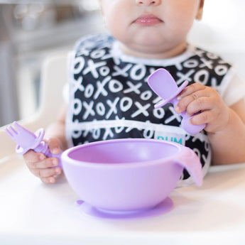 A baby sits in a high chair, wearing an XOXO patterned bib, holding Lavender Chewtensils® by Bumkins, ideal for baby-led weaning. A matching lavender bowl is placed on the tray in front of them.