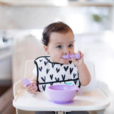 Toddler mastering baby-led weaning in a high chair using Bumkins Silicone Chewtensils®: Lavender, wearing a heart-patterned bib.